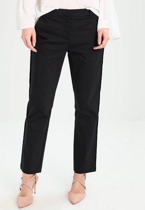 Kobieta ubrana w czarne spodnie o prostych nogawkach, białą bluzkę oraz beżowe szpilki z paskami wokół kostek, stojąca na tle jednolitej białej ściany.