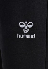 Črne športne hlače z belim vezenim logotipom z obliko čebele in besedo "hummel" spodaj. Mehka, raztegljiva tkanina.