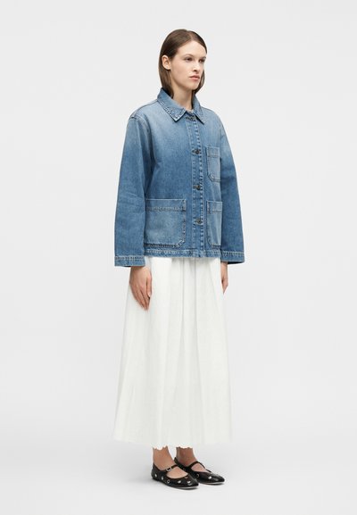 Γυναίκα που φοράει μπλε τζιν μπουφάν με μπροστινές τσέπες, μακριά λευκή φούστα και μαύρα επίπεδα πα πούτσια με λουράκια στον αστράγαλο, στέκεται μπροστά σε ένα απλό φόντο.