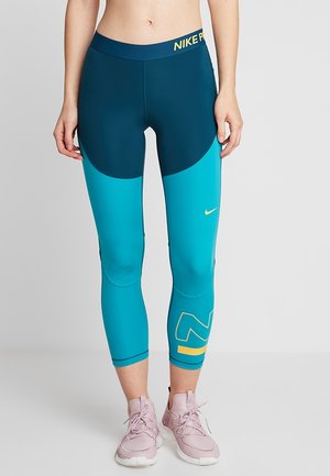 Torso e gambe femminili che indossano leggings Nike Pro blu con logo giallo e scarpe da ginnastica rosa, in piedi contro uno sfondo grigio uniforme.