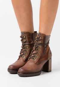 Bruine synthetische enkelboots met een gestructureerde afwerking, vetersluiting aan de voorkant, zijrits en blokhak met een patroonaccent op de kraag.