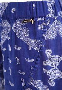 Niebieska tkanina z białym wzorem paisley, z falbanką wzdłuż krawędzi i srebrnym akcentem w postaci logo "GUESS".