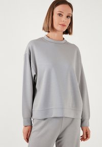 Sudadera gris claro con un cuello alto, mangas largas y un corte relajado. Está hecha de un tejido suave con una textura sutil y detalles de costura.
