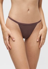 Femme portant un sous-vêtement style bikini marron avec de fines bretelles latérales, debout les mains reposant sur le haut de ses cuisses sur un fond uni.