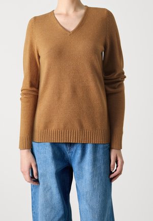 Personne portant un pull marron à col en V manches longues et un jean bleu ample sur un fond clair uni.