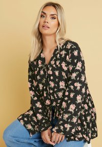 Blusa nera a fiori con rose rosa, dotata di maniche a campana lunghe, scollatura a V e un laccio in vita. Abbinata con jeans azzurri chiari.