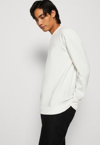 Hvid sweatshirt med rund halsudskæring, ribbede håndled og kant; har et subtilt præget logo på brystet; parret med sorte bukser.