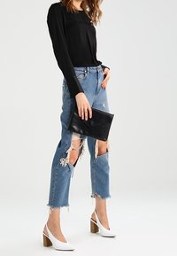 Musta pitkähihaine paita, kulutetut vaaleansiniset farkut suurilla repeämillä, musta clutch-laukku ja valkoiset kärjet korkeat korot puupohjilla.