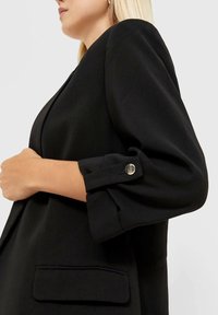 Blazer nero con una vestibilità sartoriale, spalle strutturate, polsini arrotondati con bottone a pressione e una texture elegante.