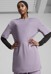 Violetinės trumpomis rankovėmis palaidinė, laisvo kirpimo, apvaliu iškirpimu ir lygia tekstūra, uždėta ant ilgos juodos marškinės. Matomi siūlių detalės.