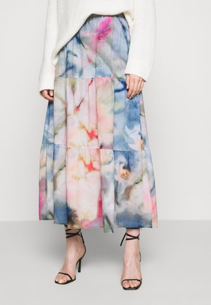 Personne portant une longue jupe à volants avec des motifs aquarelle bleus, roses et beiges, associée à des talons hauts noirs à lanières et un pull blanc.