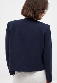 Karališkai mėlynas tekstūruotas švarkas su ilgomis rankovėmis, turintis paprastą, švarų dizainą ir tiesų apačios kirpimą, matomas iš nugaros.