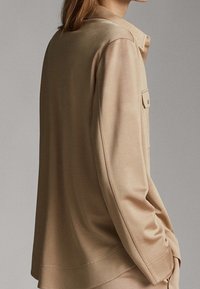 Haut beige à manches longues avec une coupe décontractée, doté d'une poche poitrine unique, d'une texture lisse et d'un large col.