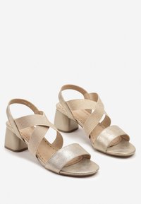 Sandales à talons blocs dorées avec des lanières croisées. Matière supérieure métallique lisse, design ouvert à l'avant, et bande de cheville texturée.