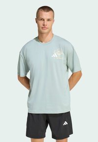 Light green short-sleeve t-shirt, soft texture, featuring a white graphic and three-stripe logo on the chest. Paired with black shorts.