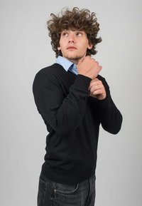 Černý pletený svetr s výstřihem do V, nošený přes světle modrou košilí s límečkem. Model pózuje s rukama, které upravují svetr, na šedém pozadí.