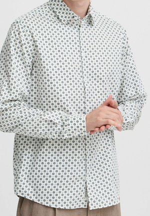Personne portant une chemise blanche à manches longues avec un petit motif noir en forme de rayons de soleil, mains jointes au niveau de la taille.