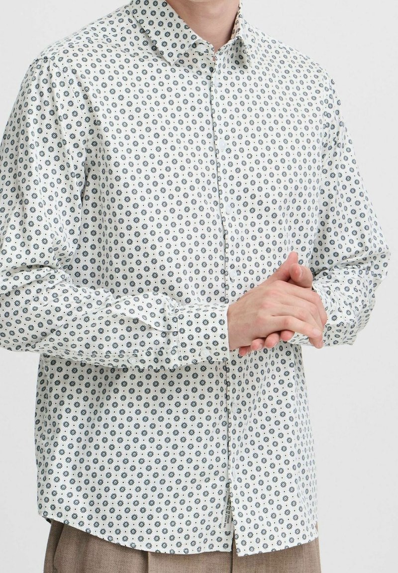 Personne portant une chemise blanche à manches longues avec un petit motif noir en forme de rayons de soleil, mains jointes au niveau de la taille.