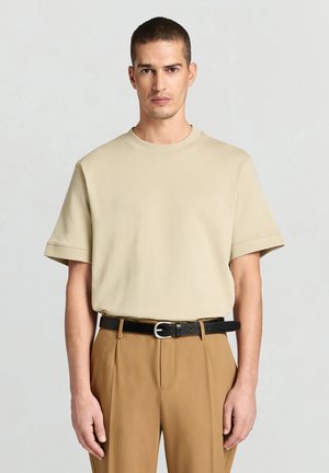 Beiges T-Shirt mit kurzen Ärmeln und rundem Ausschnitt, kombiniert mit hellbraunen Hosen und einem schwarzen Gürtel mit silberner Schnalle.