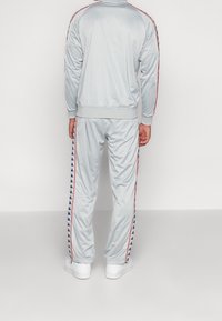 Homme portant un survêtement gris clair avec des bandes rouges et bleues du logo sur les manches et les jambes du pantalon, debout devant un fond blanc.