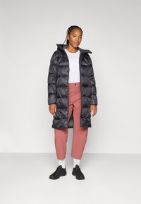 Mlada žena stoji u streetwear odjeći koja se sastoji od dugog crnog puffer kaputa, bijele majice, ružičastih hlača i crnih tenisica, nasuprot neutralnoj pozadini.