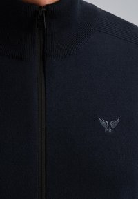 Tamno plavi zip-up pulover s rebrastim ovratnikom, sa crnim patentnim zatvaračem i malim sivim logotipom na lijevoj strani prsnog koša. Glatka tekstura.
