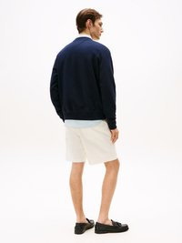 Mørkeblå sweatshirt med rund hals, avslappet passform, brukt over en lyseblå skjorte. Kombinert med hvite shorts og svarte loafers.