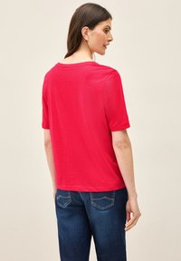 Rød kortærmet T-shirt med afslappet pasform og rund halsudskæring. Materialet virker glat; kombineret med mørkeblå jeans med baglommer.