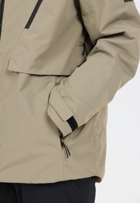 Beige buitenjack met een rits aan de voorkant, grote borstzak, verstelbare manchetten en een trekkoord aan de onderkant. Gemaakt van duurzaam, waterafstotend materiaal.
