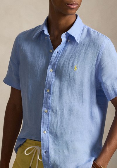 Άνδρας που φοράει ανοιχτό μπλε πουκάμισο με κοντά μανίκια και κίτρινο λογότυπο, καθώς και κίτρινο παντελόνι με λευκό κορδόνι.