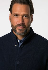 Mörkblå texturerad jacka med hög krage, tryckknappar och ett fint stickat mönster, lagd över en blå skjorta med prickar.