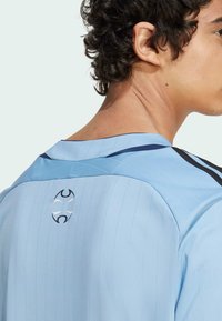 Light blue sports jersey with pinstripes, featuring a black and navy collar, and a circular logo in black on the back. Soft fabric texture.