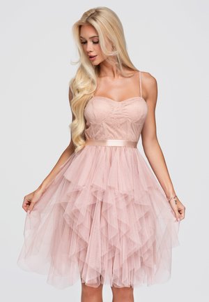Femme blonde portant une robe bustier rose pâle avec une jupe en tulle superposée, regardant vers le bas tout en tenant les côtés de la jupe.