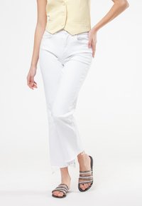 Jean flare blancs avec un ourlet effiloché, associés �à des sandales noires à brides scintillantes. Le haut est un gilet jaune clair.