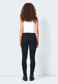 Jeans negros ajustados con cintura alta, dos bolsillos traseros y una textura suave, combinados con una camiseta de tirantes blanca de canalé y zapatillas negras.