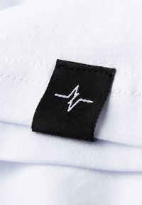 Schwarzes Stoffetikett mit weißem Faden, das ein Herzschlag-Design zeigt, befestigt an einem gefalteten weißen Textil.