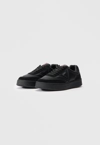 Une paire de baskets basses noires avec des semelles épaisses et texturées, des lacets sur le devant, et un logo de la marque sur le côté et la languette.