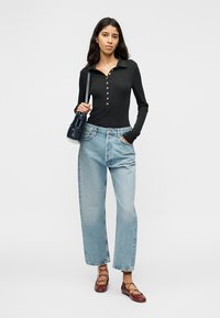 Μαύρο πουκάμισο με μακριά μανίκια και κουμπιά, ανοιχτό μπλε τζιν ψηλόμεσο, καφέ δερμάτινα παπούτσια με κορδόνια, κρατώντας μια μικρή μπλε σκούρα τσάντα. Απλό φόντο.