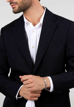 Blazer blu navy con una vestibilità slim, abbinato a una camicia bianca con bottoni. Bracciale argento al polso, che mostra una texture a rete e un dettaglio della chiusura liscia.