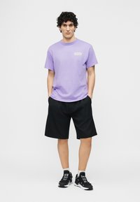 Νεαρός με σγουρά μαλλιά στέκεται με τα χέρια στις τσέπες φορώντας μωβ μπλουζάκι, μαύρα σορτς, άσπρες κάλτσες και μαύρα αθλητικά παπούτσια.