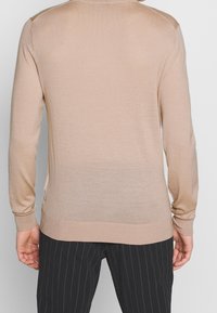 Pull en tricot beige à manches longues et à poignets côtelés. Présente des fentes sur les côtés et un dégradé subtil de couleur allant d'un beige clair à un beige plus foncé.