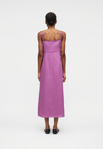 Femme aux cheveux tressés portant une robe violette sans manches, longue jusqu'aux chevilles, avec des bretelles à nouer, et des chaussures noires sans lacets, debout de dos.