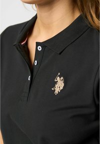 Zwarte polo met een gestructureerde stof, voorzien van een drieknopenlijst, contrasterende kraag en een lichtgekleurde geborduurde logo.