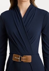 Vestito avvolgente blu navy con maniche lunghe, dotato di una cintura in finta pelle marrone con una fibbia argentata e dettagli di cucitura. Tessuto liscio e strutturato.