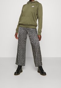 Oliwkowa zielona bluza z kapturem z kieszenią z przodu i białym logo, zestawiona z szerokimi spodniami w czarno-biały wzór oraz czarnymi sznurowanymi botkami.