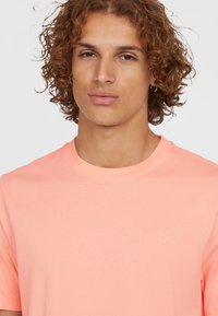 Lichtroze t-shirt met korte mouwen, gemaakt van katoen, met een ronde hals en een subtiele geborduurde logo op de borst. Glad textuur.