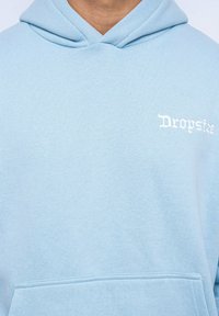 Helle blaue Kapuzenjacke aus weichem Stoff mit einer Kängurutasche und "Dropsize" in Weiß auf der linken Bruststickerei.