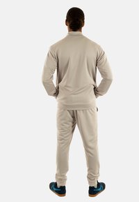 Tuta di colore beige chiaro con colletto zip e polsini elastici, abbinata a scarpe da ginnastica blu scuro con accenti azzurri.