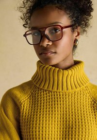 Frau mit lockigem Haar, die große braune Brille, goldene Tropfenohrringe und einen senfgelben Strickrollkragenpullover trägt, vor einem beigen Hintergrund.