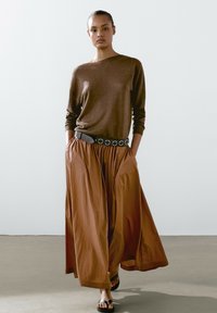 Hnědý dlouhý rukáv top spárovaný s sukní širokého střihu v měděné barvě. Obsahuje černý pásek se stříbrnými kulatými akcenty. Hladká tkanina a uvolněný střih.